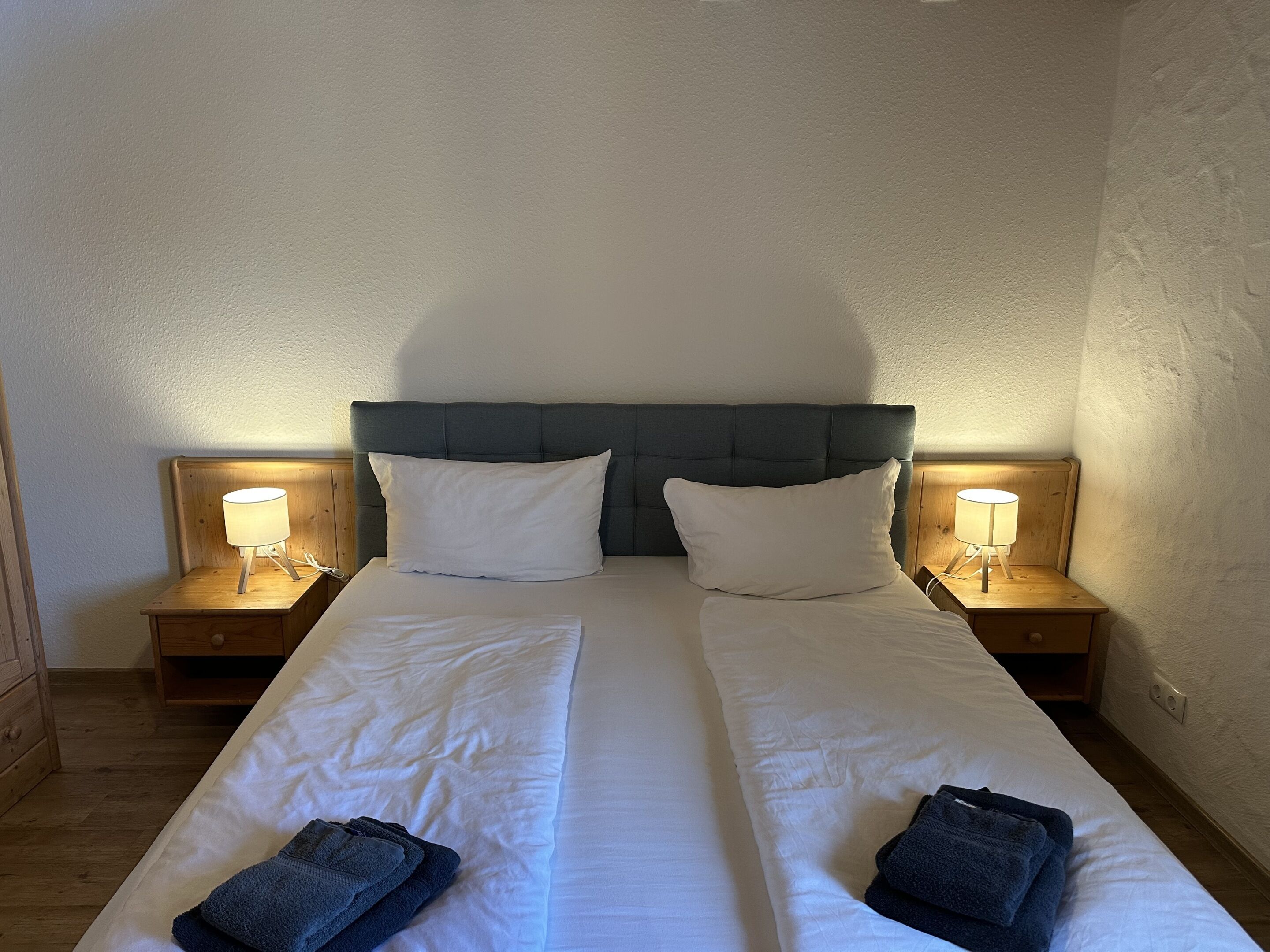 A double bed with two pillows, two bedside tables with lamps, and folded towels on the bed.