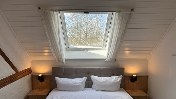 Ein Schlafzimmer mit Oberlicht, zwei Nachttischen, einem Bett mit weißer Bettwäsche und Blick auf Bäume.