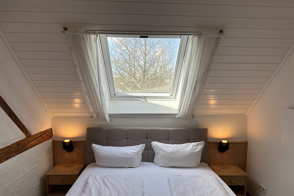 A bedroom with a skylight, two bedside tables, a bed with white bedding, and a view of trees outside.