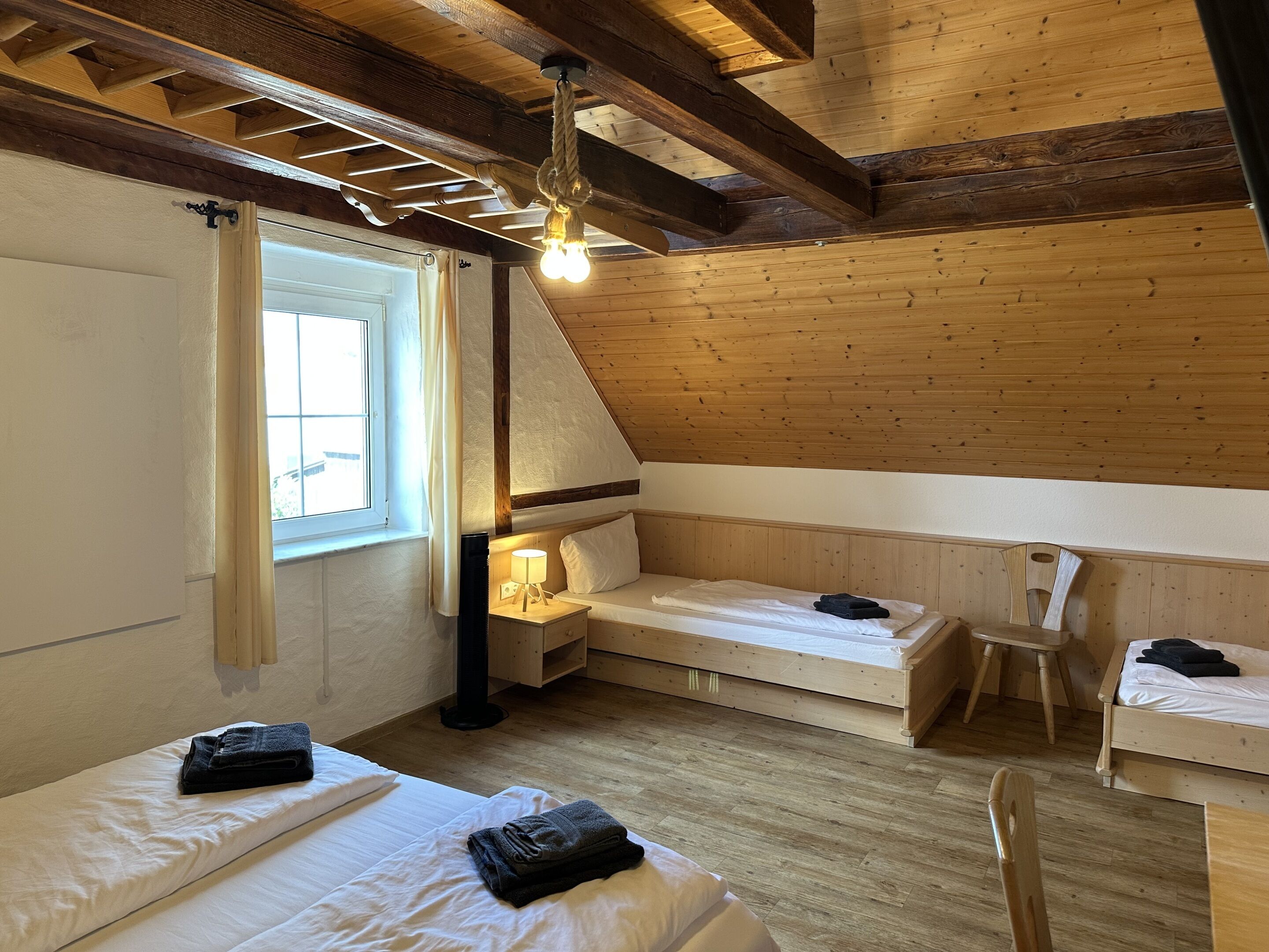 A cozy attic bedroom with wooden beams, a sloped ceiling, and a window with curtains. There are two beds, a chair, and a bedside table with a lamp.