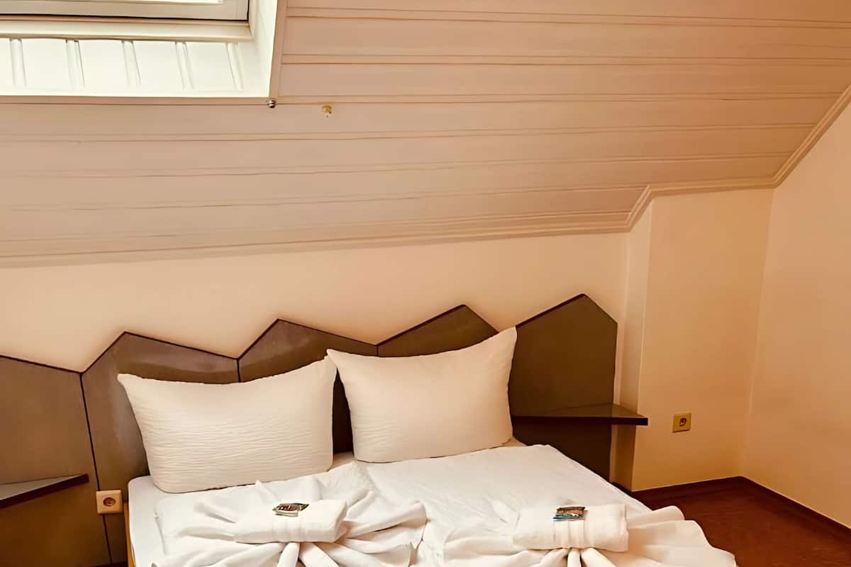 A neatly made bed with pillows and a folded towel on a wooden bed frame in a room with a sloped ceiling and a window.