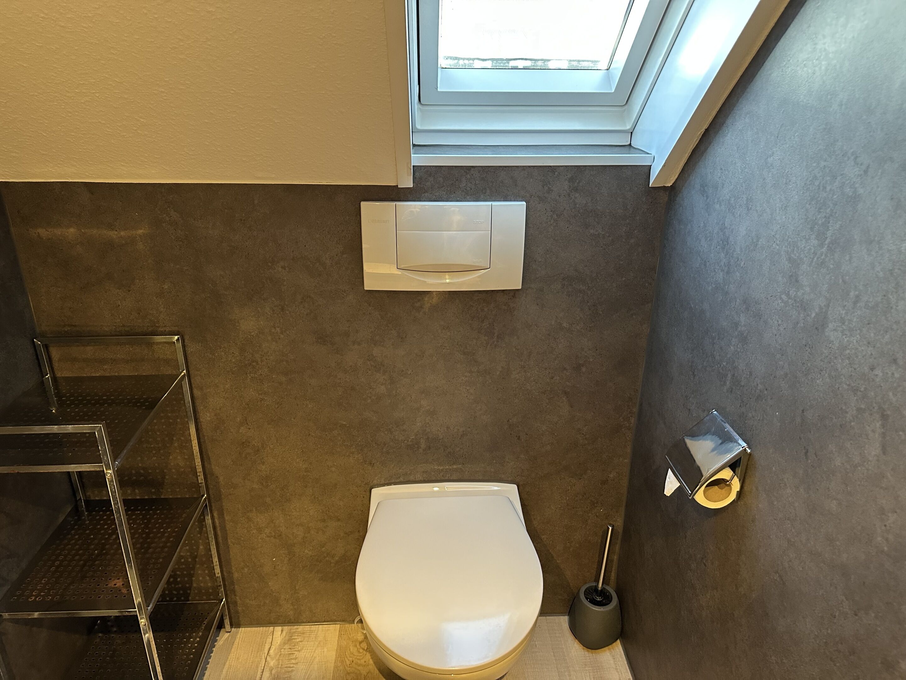 Een kleine badkamer met toilet, een metalen plank, een toiletborstelhouder en een raam boven het toilet.