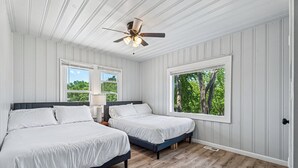 Ein Schlafzimmer mit zwei Betten, einem Deckenventilator und großen Fenstern mit Blick auf Bäume.