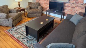 Una sala de estar con pared de ladrillo, un televisor de pantalla plana, dos sillones estampados, un sofá oscuro y una mesa de centro.