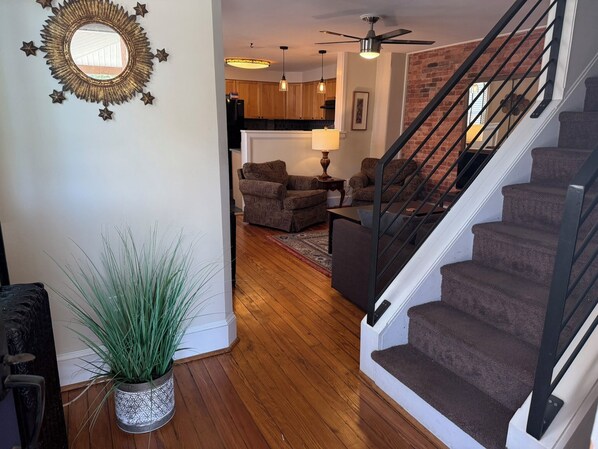 A spacious living area with a wooden floor, a staircase with a black railing, and a kitchen in the background.
