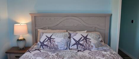 Cama com cabeceira, duas almofadas com estampas de estrelas-do-mar e corais, mesa de cabeceira com abajur e parede azul.