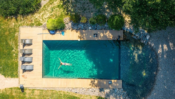 Vue aérienne d’une piscine rectangulaire dotée d’une terrasse en bois, entourée de verdure et bordée d’un chemin de galets.