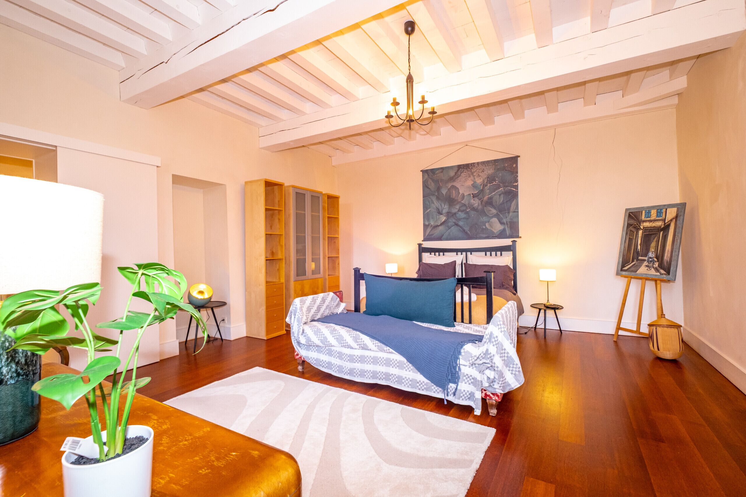 A bedroom with a bed, bedside tables, a chandelier, a painting, and a potted plant.