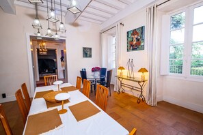 Et spiseområde med et langt bord dækket til fire personer, omgivet af træstole. Der hænger lysarmaturer over bordet, et maleri på væggen og et vindue med gardiner.