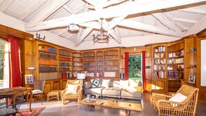 Une bibliothèque spacieuse, dotée de hauts plafonds et de poutres apparentes, comprenant un grand canapé, des fauteuils en osier et de nombreuses étagères remplies de livres.