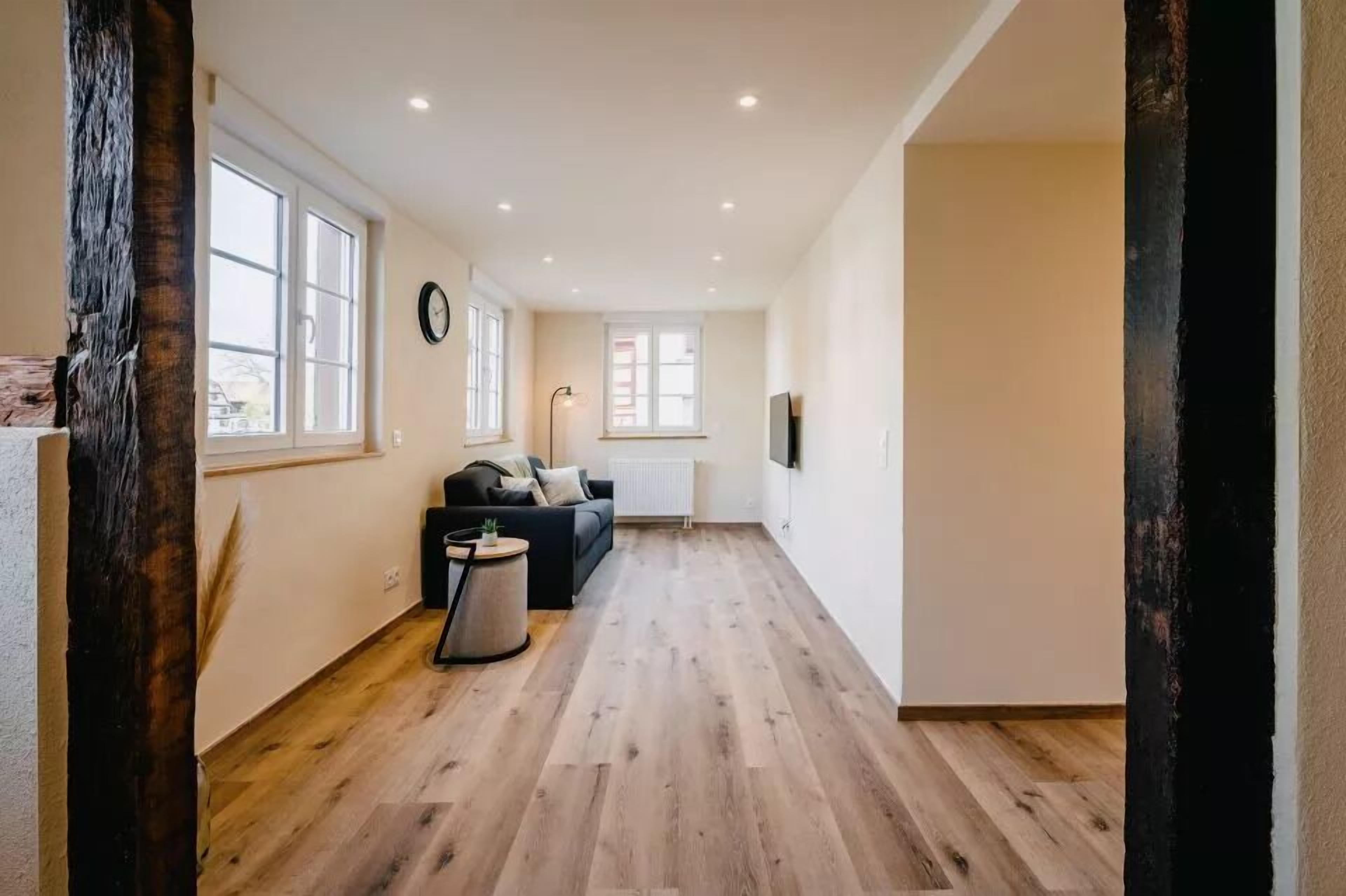 A spacious living area with a wooden floor, a black sofa, a round coffee table, and a large window providing natural light.
