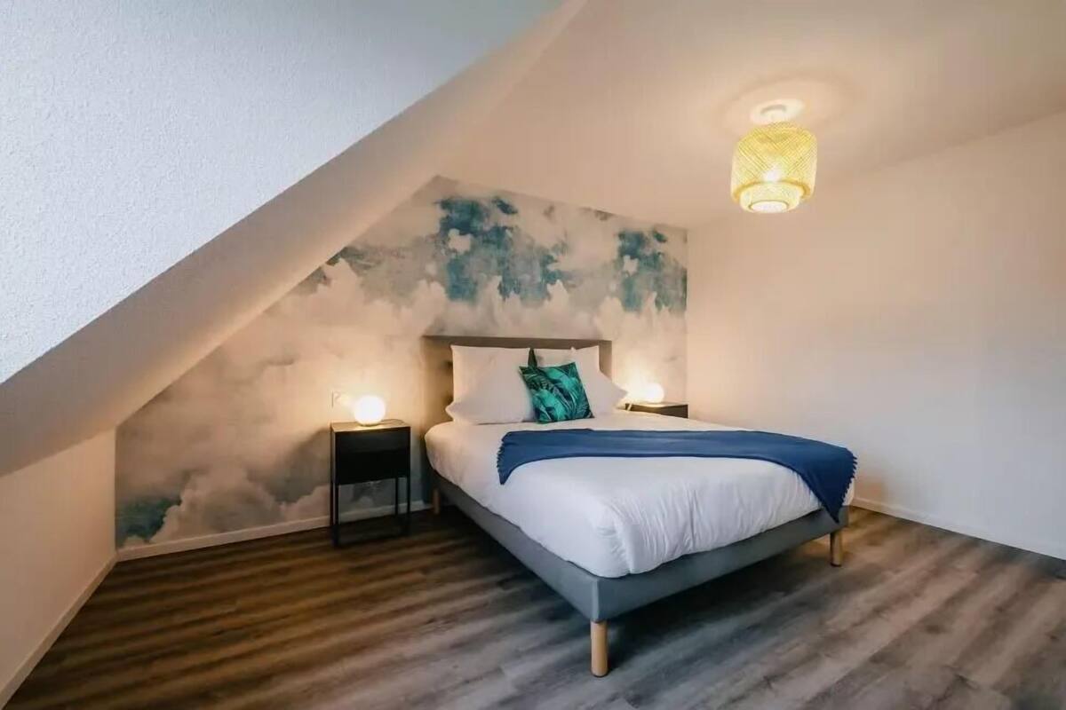Un dormitorio con techo inclinado, una cama con ropa de cama blanca y una manta azul, dos lámparas de noche y un mural en la pared.