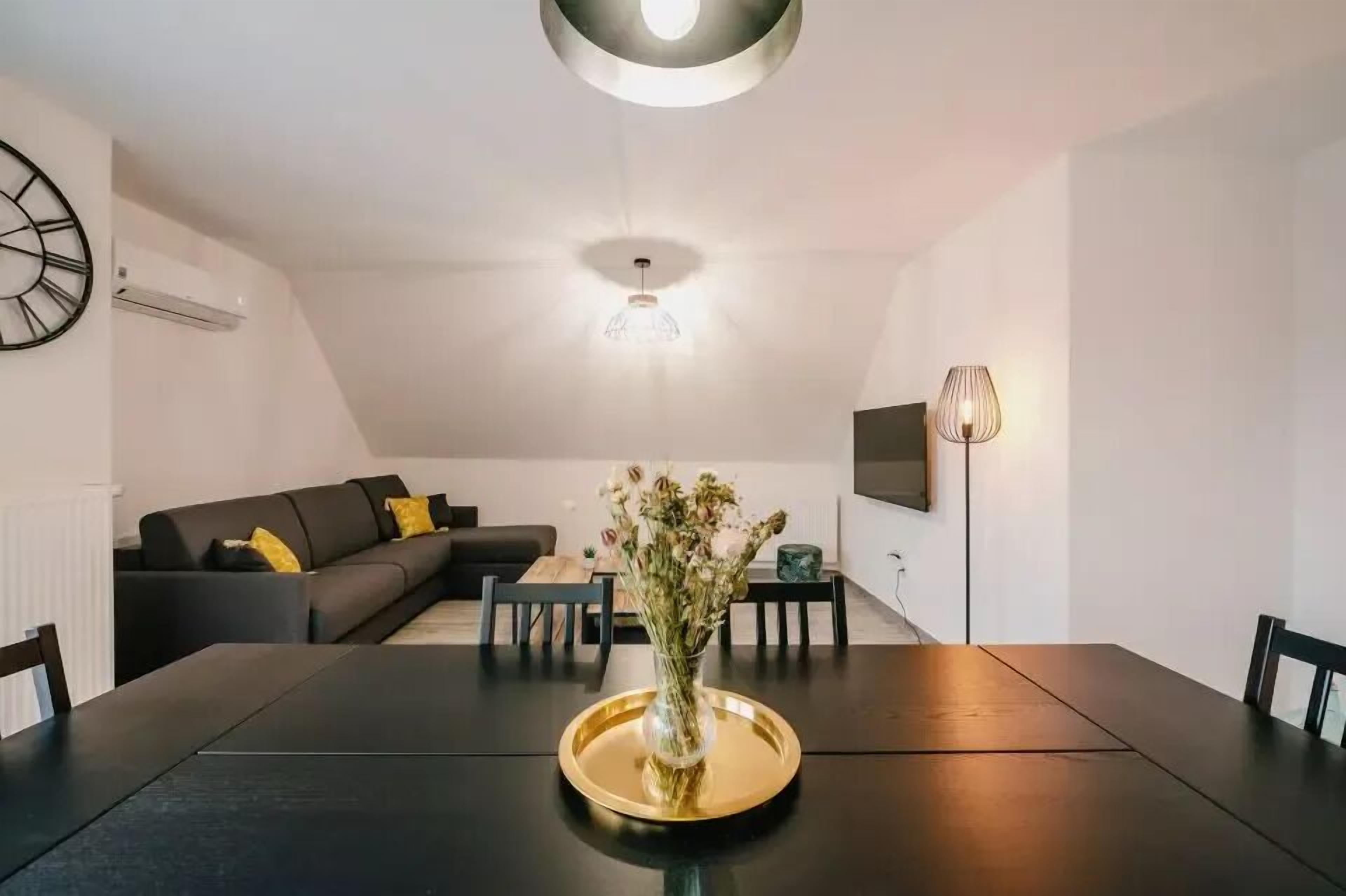 Ein modernes Wohnzimmer mit einem dunkelgrauen Sofa, einem Esstisch mit Blumen und einer Deckenleuchte.