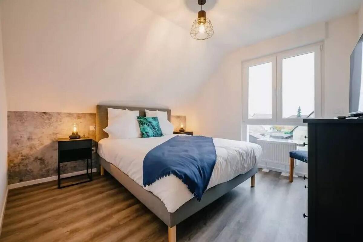 Un dormitorio con techo inclinado, una cama con ropa blanca y un plaid azul, una mesita de noche con una lámpara y vistas al exterior a través de una ventana.