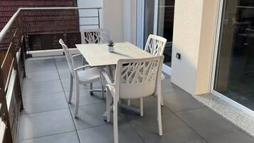 A balcony with a white table and chairs, a small potted plant, and a sliding glass door.