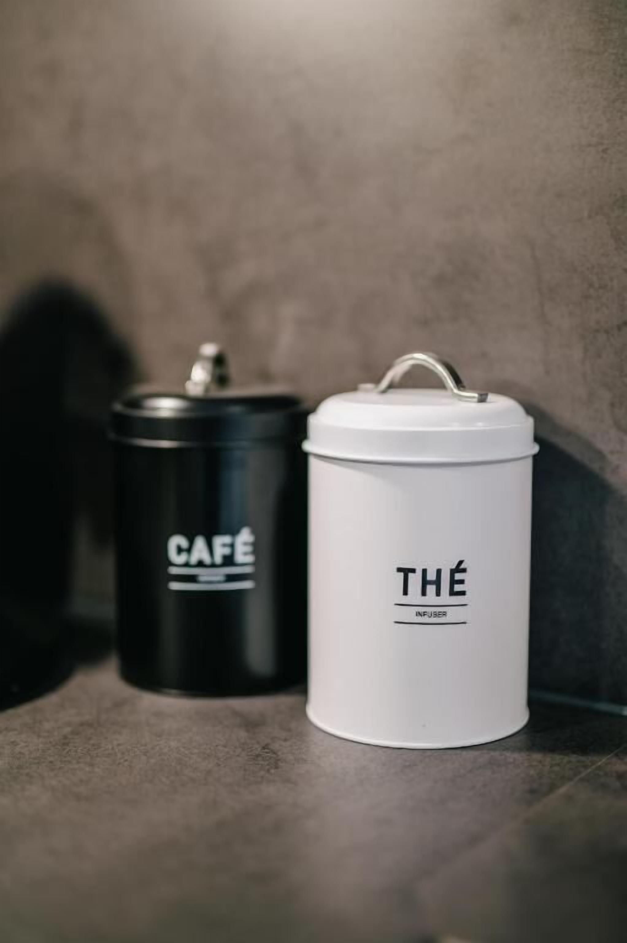 Two containers, one black and one white, labeled 'CAFÉ' and 'THÉ' respectively, placed on a tiled surface.