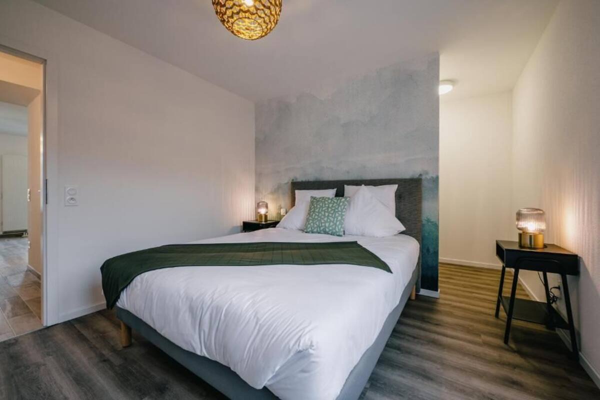 Un dormitorio con una cama grande, dos mesitas de noche con lámparas, un cabecero texturizado y una puerta que da a otra habitación.