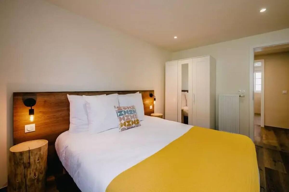 Un dormitorio con cabecera de madera, cama con ropa de cama blanca y un plaid amarillo, dos mesitas de noche con lámparas de pared, y un armario.
