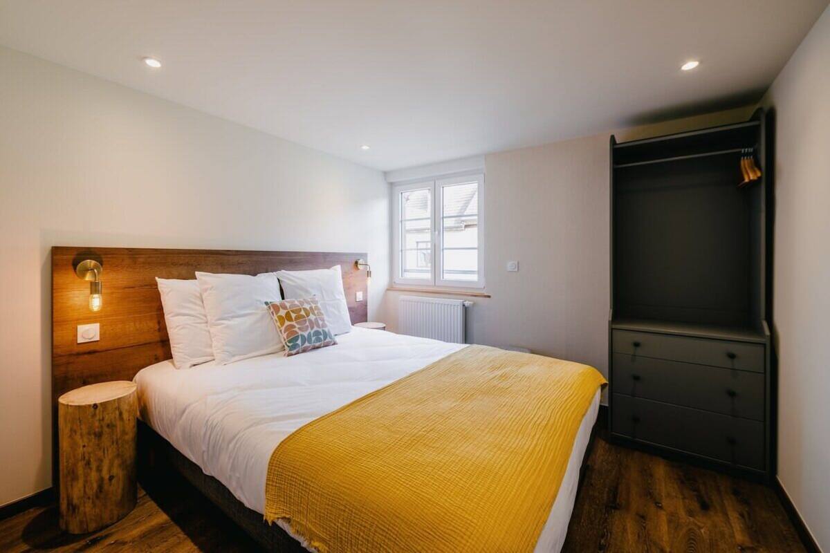 Un dormitorio con cabecera de madera, cama con ropa de cama blanca y una manta amarilla, mesita de noche, radiador y armario.