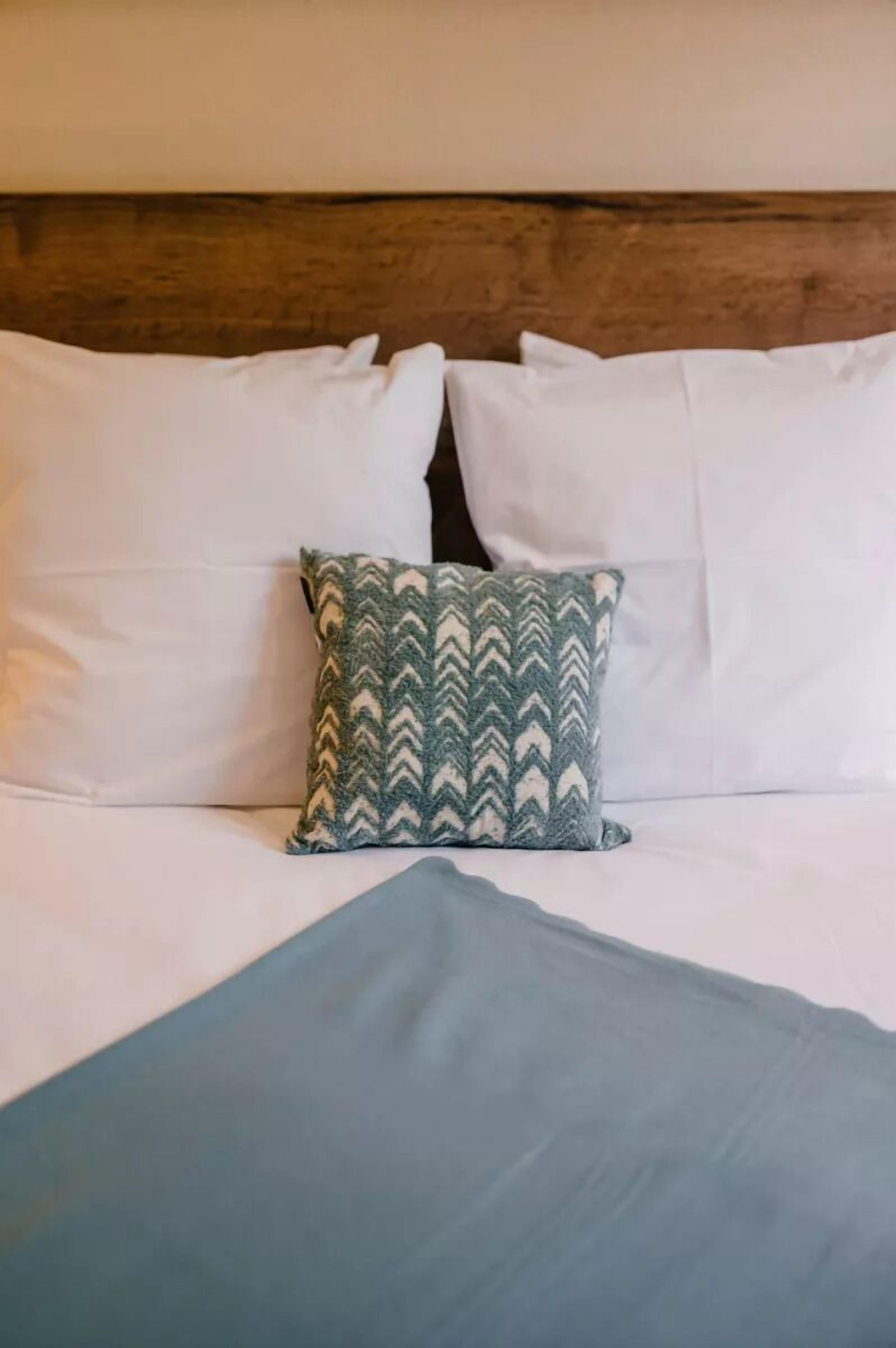 Ein Bett mit weißen Kissen, einem gemusterten Kopfkissen und einer blauen Decke. Dazu ein Holz-Kopfteil.