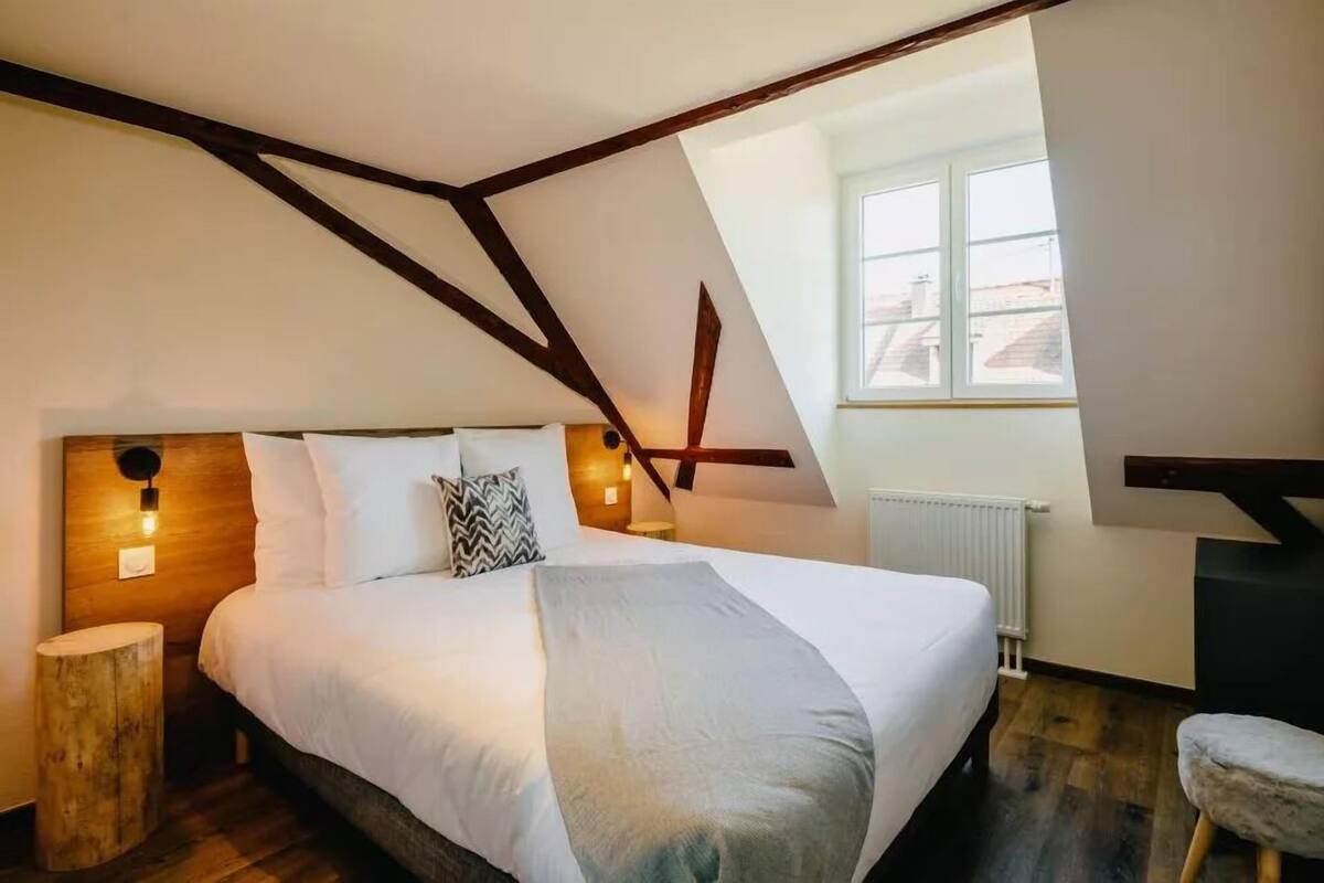 Un dormitorio con techo inclinado, una cama con ropa de cama blanca, una mesita de noche de madera, un taburete pequeño y una ventana con vista a edificios.
