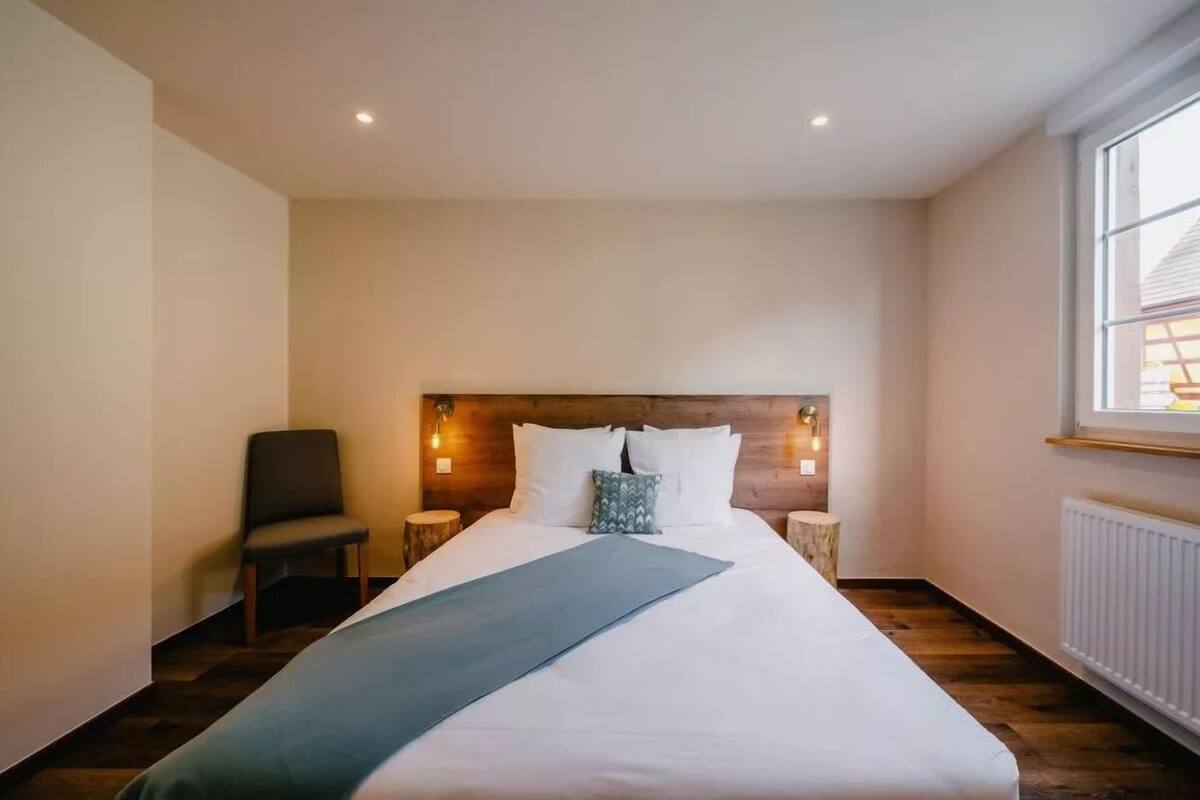 Habitación de hotel con una cama grande, una silla, mesitas de noche y un ventanal con vistas.