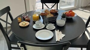 Ruokapöytä on katettu aamiaistarvikkeilla, kuten croissantteja, hedelmiä ja juomia. Huoneesta on lasioven kautta näkymä ympäröiviin rakennuksiin.