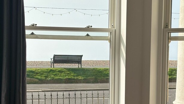Una habitación con una mesa redonda y cuatro sillas, una ventana que ofrece vistas a un banco y una zona verde, y una guirnalda de luces.
