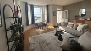 Una sala de estar moderna con un sofá, una mesa de comedor y sillas. Hay un ventanal con cortinas, un espejo en la pared y una planta sobre un pedestal.