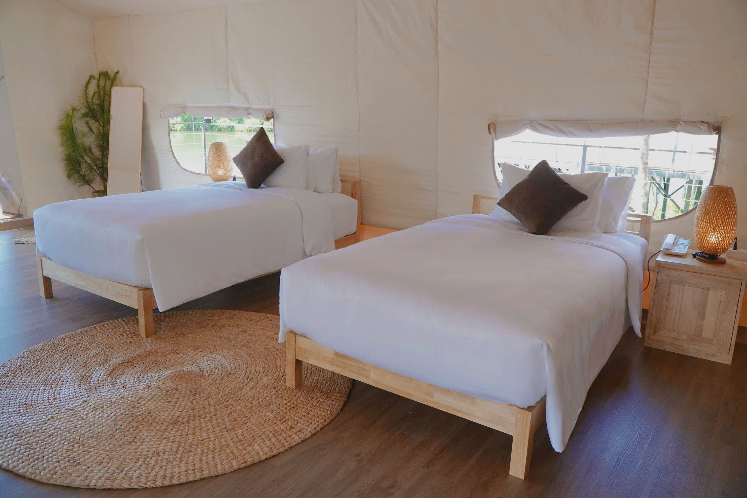 Two single beds with wooden frames and white bedding, a woven rug, a small wooden nightstand with a lamp, and a window with a view of greenery.