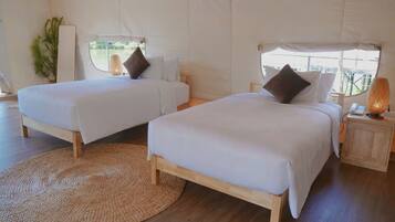 Two single beds with wooden frames and white bedding, a woven rug, a small wooden nightstand with a lamp, and a window with a view of greenery.