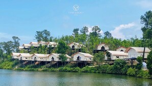 A series of elevated, tent-like structures with multiple levels, surrounded by lush greenery and a body of water. - La Vela Legacy Resort (Hue)