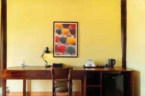 A hotel room with a wooden desk, a chair, a desk lamp, a framed artwork, a refrigerator, and a coffee maker. - La Vela Legacy Resort (Hue)