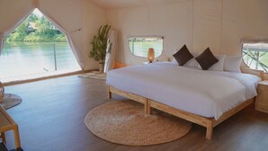 A bedroom with a large bed, a round woven rug, a nightstand, a lamp, and a window with a view of greenery and a building. - La Vela Legacy Resort (Hue)