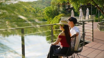 Una pareja sentada en un banco con vista a un lago, con un puente y árboles al fondo.