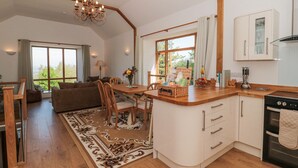 A spacious kitchen with a large island, wooden cabinets, and a built-in oven. There is a dining area with a table and chairs, and a view of greenery through large windows. - Newlands Barn (Wellington)