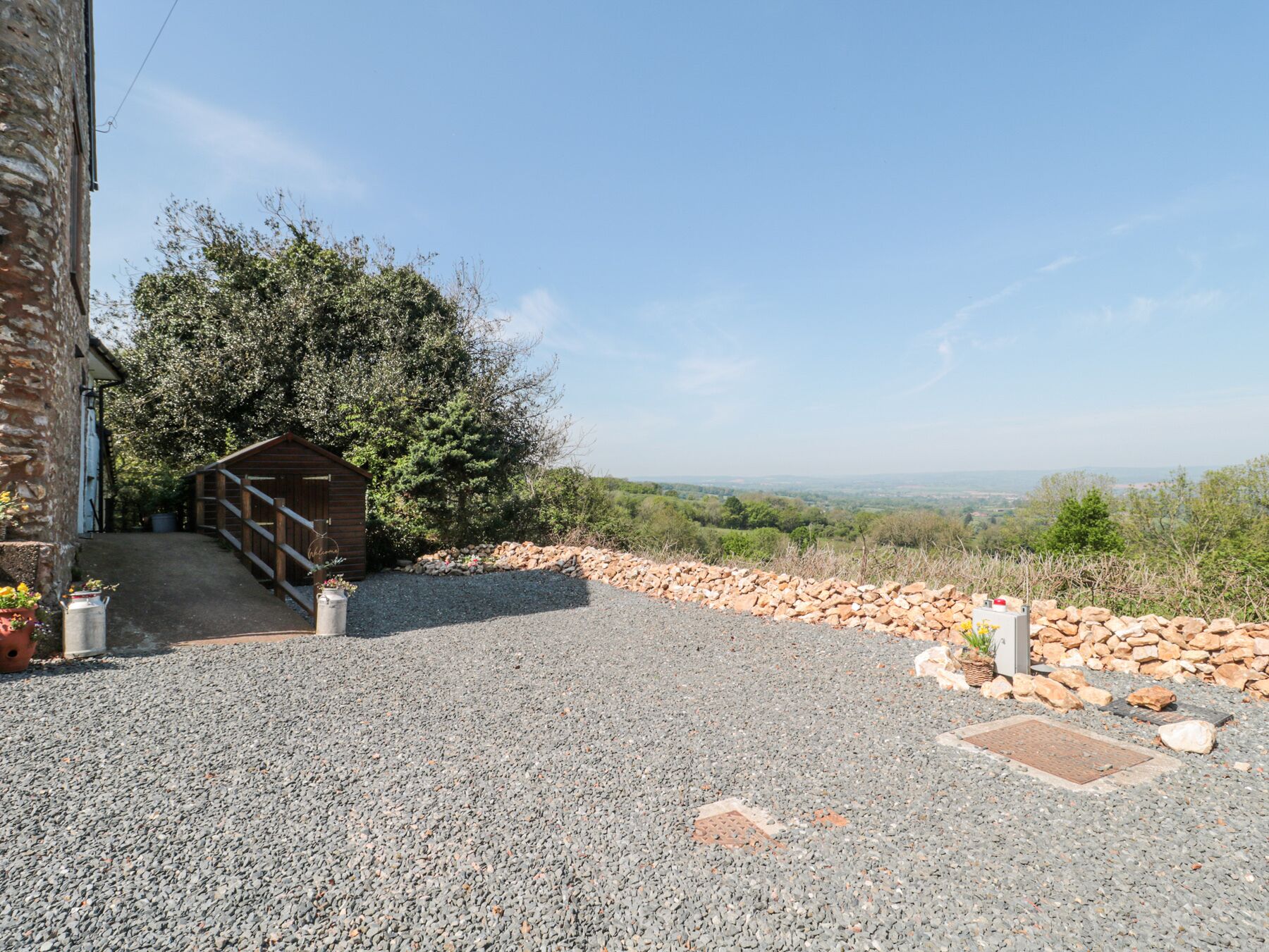 Ein Schotterweg führt zu einem Holzbauten mit einer Steinmauer und Blick auf die Landschaft.