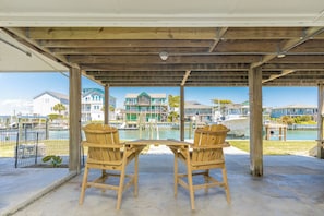 A covered outdoor seating area with two wooden chairs and a table, overlooking a marina with boats and buildings.