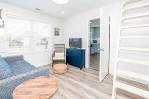 A room with a blue sofa, a wooden coffee table, a wicker ottoman, a television on a blue cabinet, and a view into a kitchen area.