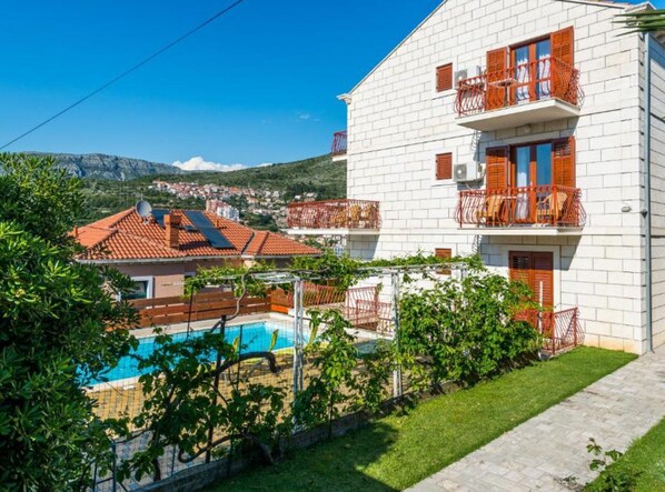 A white building with balconies, a swimming pool, and a view of a town and mountains.