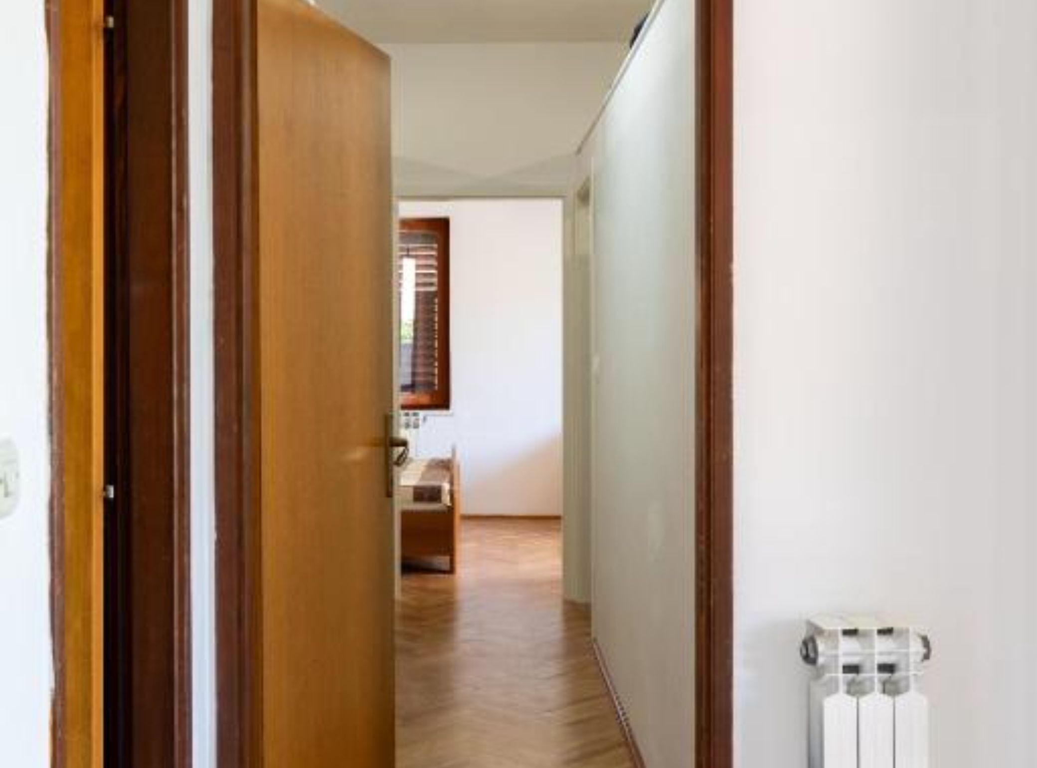 Un couloir avec une porte en bois, un radiateur et une fenêtre avec des stores.