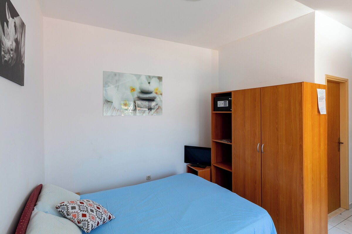 A bedroom with a bed, a wooden wardrobe, a television, and a painting on the wall.