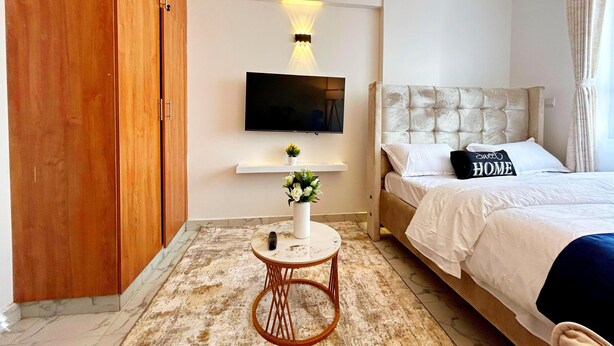 Een moderne slaapkamer met een groot bed, een aan de muur bevestigde televisie, een klein rond tafeltje met een vaas en een houten garderobe.