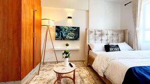Een moderne slaapkamer met een bed, een aan de muur bevestigde televisie, een vloerlamp en een klein tafeltje met een vaas.