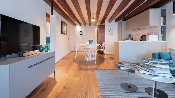 Een moderne woonkamer met een houten plafond, een flatscreen televisie, een eetgedeelte met witte stoelen en een keuken met een magnetron en boeken.