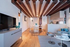 A modern living room with a wooden ceiling, a flat-screen TV, a dining area with white chairs, and a kitchen area with a microwave and books. - Ca' Grifalconi (Venice)