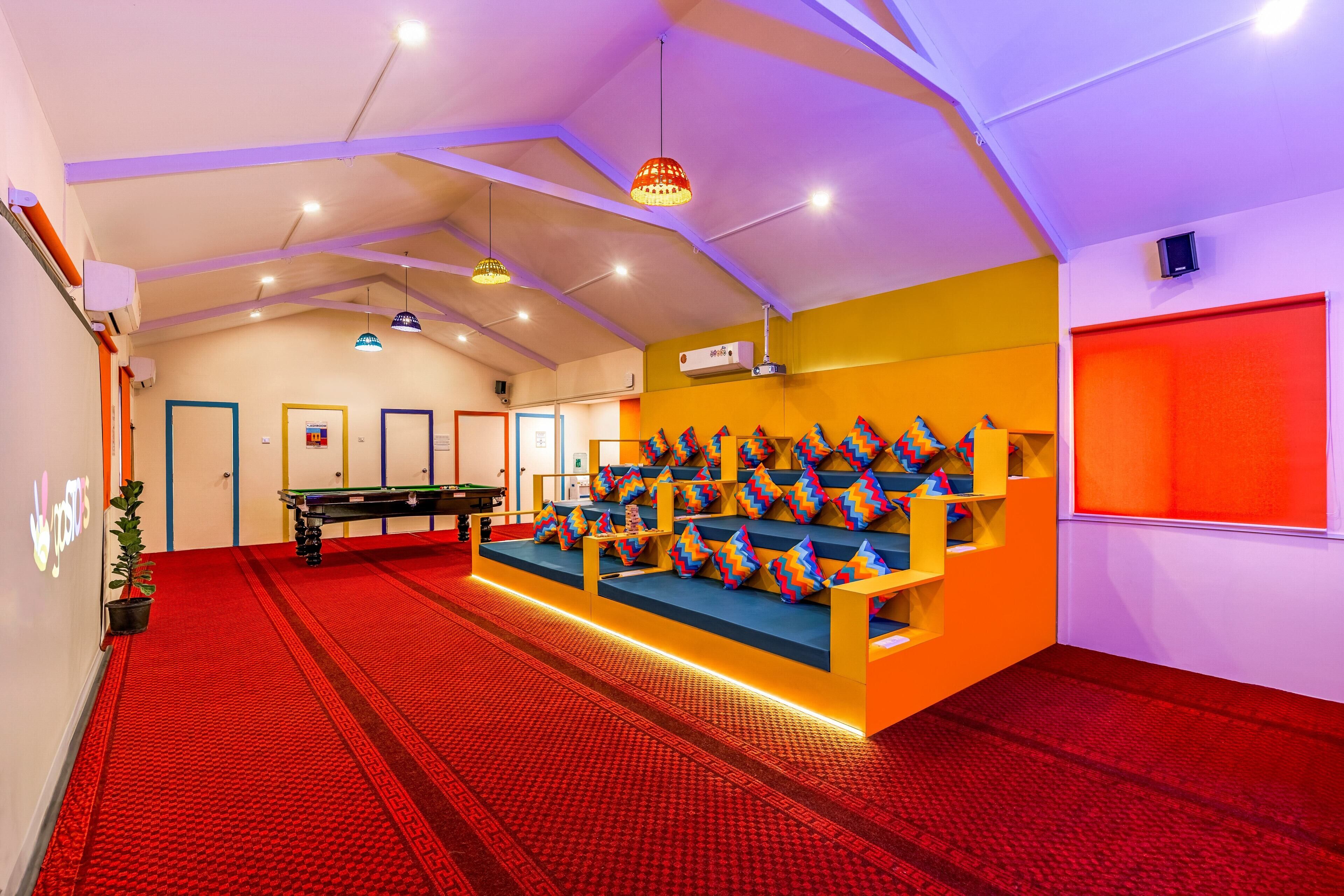 A room with a pool table, a seating area with colorful cushions, and a red carpet.