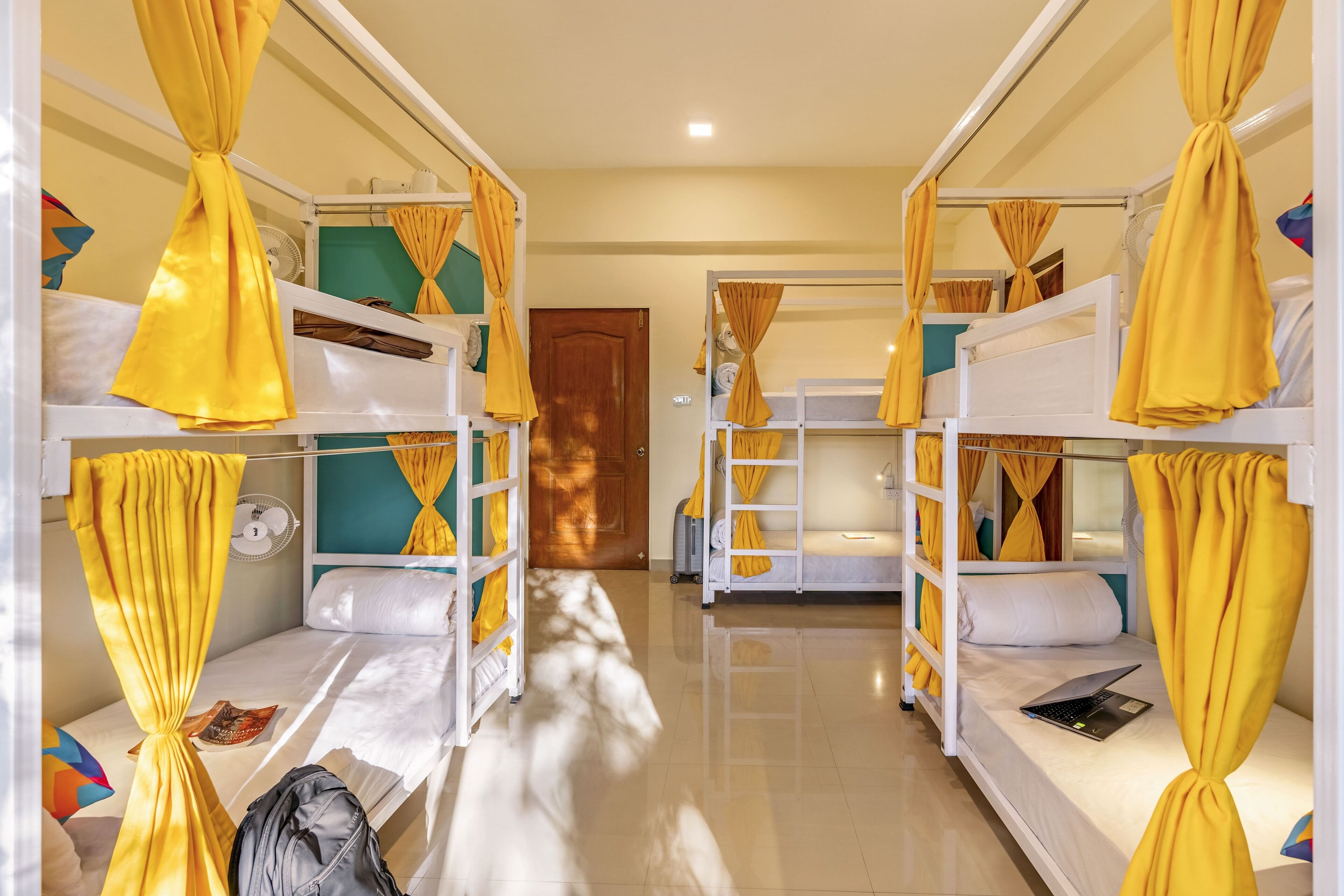 Una habitación con literas, cada una con cortinas amarillas atadas, un ventilador y un ordenador portátil sobre una de las camas.