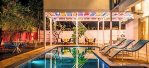 A pool area with lounge chairs and tables, surrounded by a covered patio with colorful ceiling lights. - goSTOPS Alibaug (Alibag)