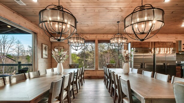 A wooden dining area with a long table and chairs, large windows, and a modern kitchen with stainless steel appliances.